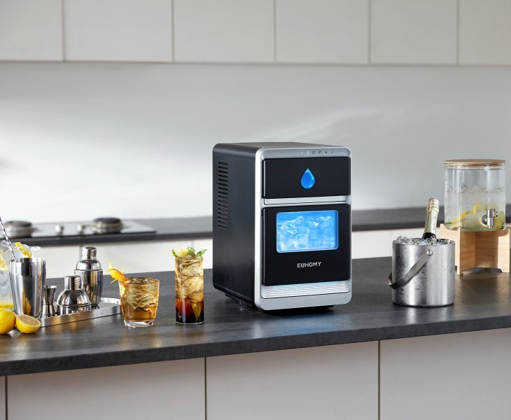 A countertop ice maker placed next to a glass of iced water on a counter.