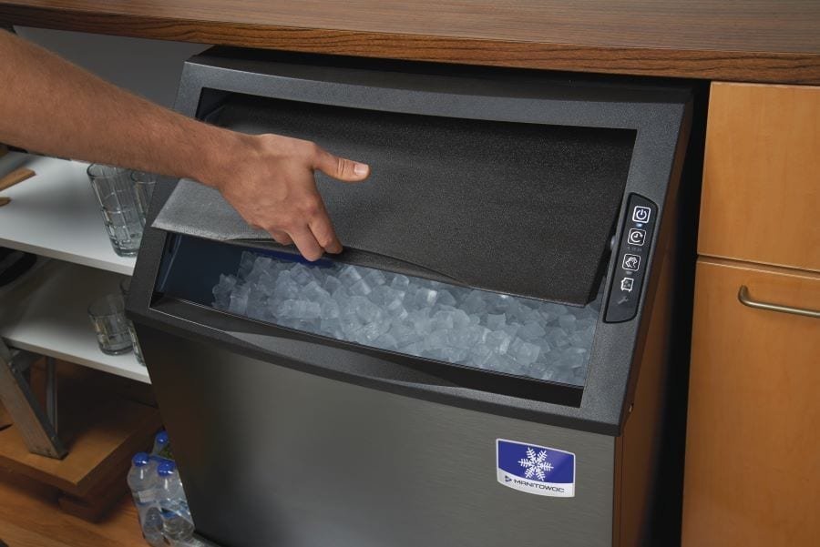 An open undercounter ice maker sits in a clean kitchen near a glass of water.